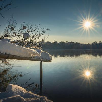 Ein Handlauf ist mit viel Schnee bedeckt. Der Sonnenuntergang spiegelt sich im See.