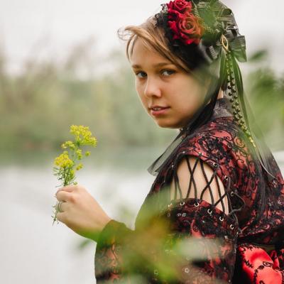 Eine junge Frau in einem roten Kleid mit Rosen im Haar hält eine gelbe Blume in der Hand und blickt überrascht über ihre Schulter.