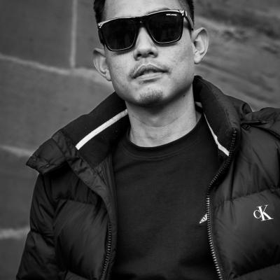 Ein junger Mann asiatischer Herkunft sitzt cool und gelassen auf einer Treppe, trägt eine Sonnenbrille und eine Winterjacke. Im Hintergrund ist eine leicht verschwommene Mauer zu sehen.